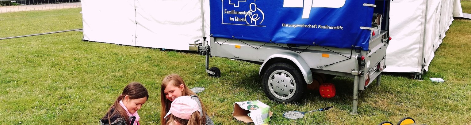 Spielmobil Ein Auto-Anhänger mit Aufschrift - Förderverein Haus der Familie - und dem Förderhinweis Diakoniegemeinschaft Paulinenstift - steht auf einer Wiese, davor spielende Kinder.