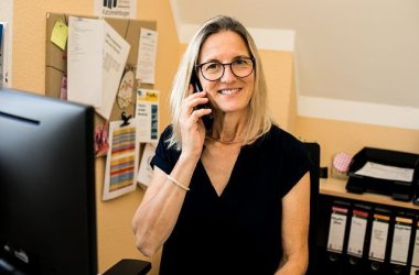 Eine Frau mit halblangen blonden Haaren telefoniert an ihrem Schreibtisch.
