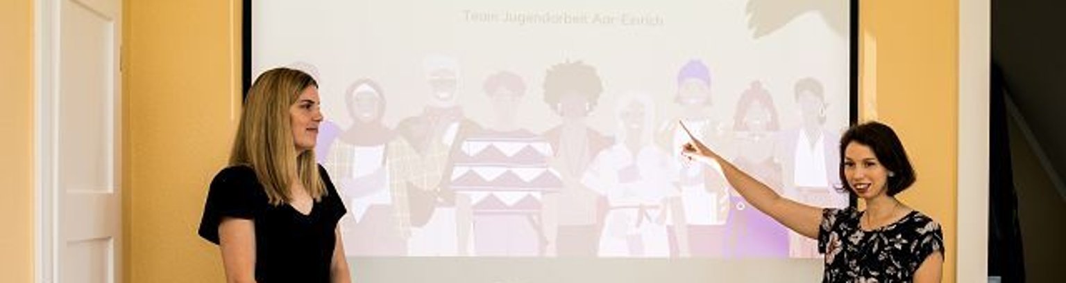Ehrenamtsschulung Zwei junge Frauen stehen vor einer Leinwand mit gezeichneten Menschen. Eine zeigt darauf.