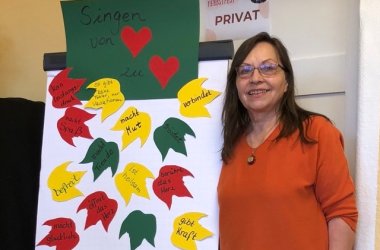 Eine dunkelhaarige Frau steht neben einem Plakat mit aufgeklebten Herbstblättern aus Papier auf denen gute Argumente für gemeinsames Singen aufgeschrieben wurden