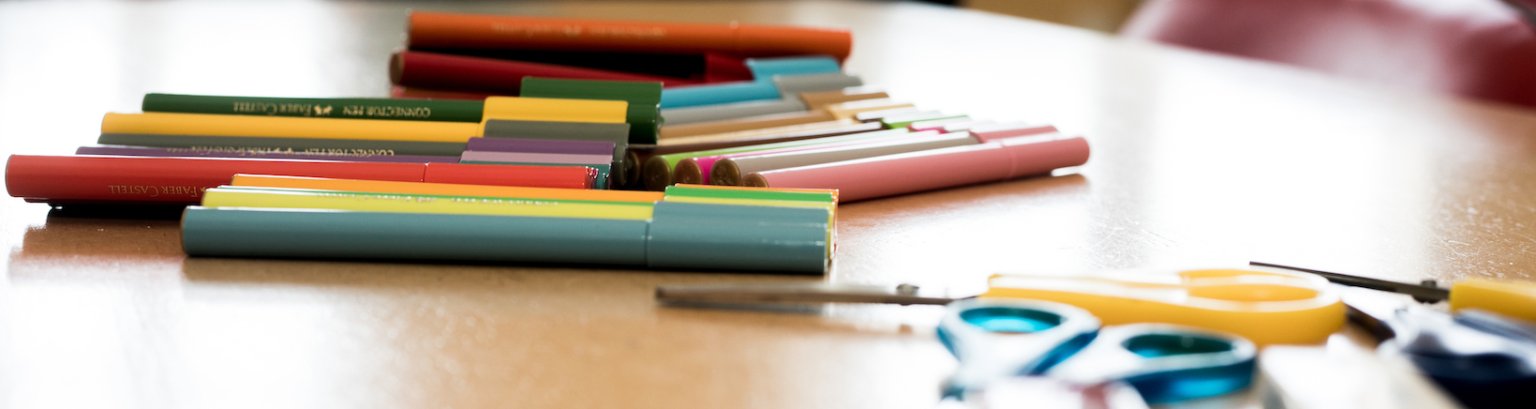 Bildausschnitt: Nahaufnahme. Auf einer Tischplatte liegen Filzstifte und Bastelscheren
