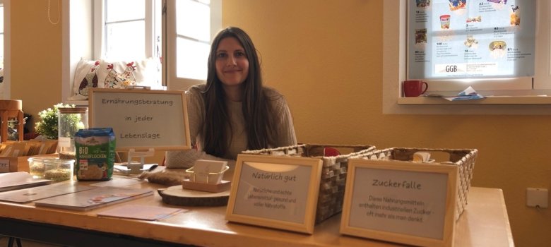 Eine junge Frau mit langen dunklen Haaren sitzt hinter einem Tisch auf dem Informationsmaterial zu guten und schlechten Nahrungsmitteln aufgebaut sind.