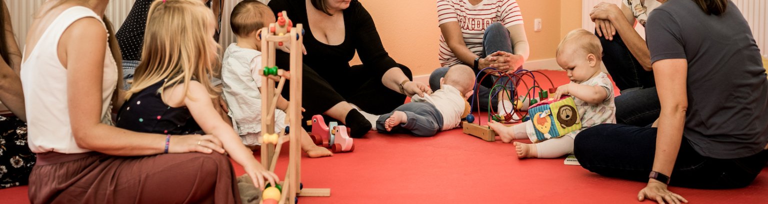 Bildausschnitt: Mütter sitzen auf dem Boden, ihre Köpfe sind nicht zu sehen. Ihre Kleinkinder spielen vor ihnen.