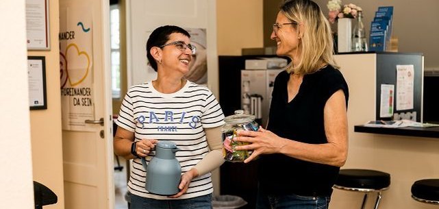 Eine Frau mit längeren blonden Haaren und eine  Frau mit kurzen dunklen Haaren stehen beieinander und lächeln sich an. Eine hält eine KaffeeKanne in der Hand, die andere ein Glas mit Teebeuteln.