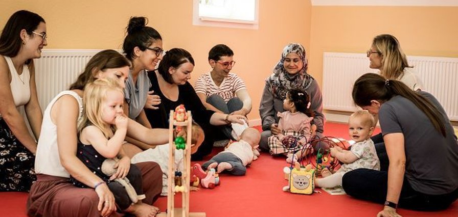 Mütter sitzen mit Ihren spielenden Kleinkindern auf dem Boden und beobachten das Spiel.