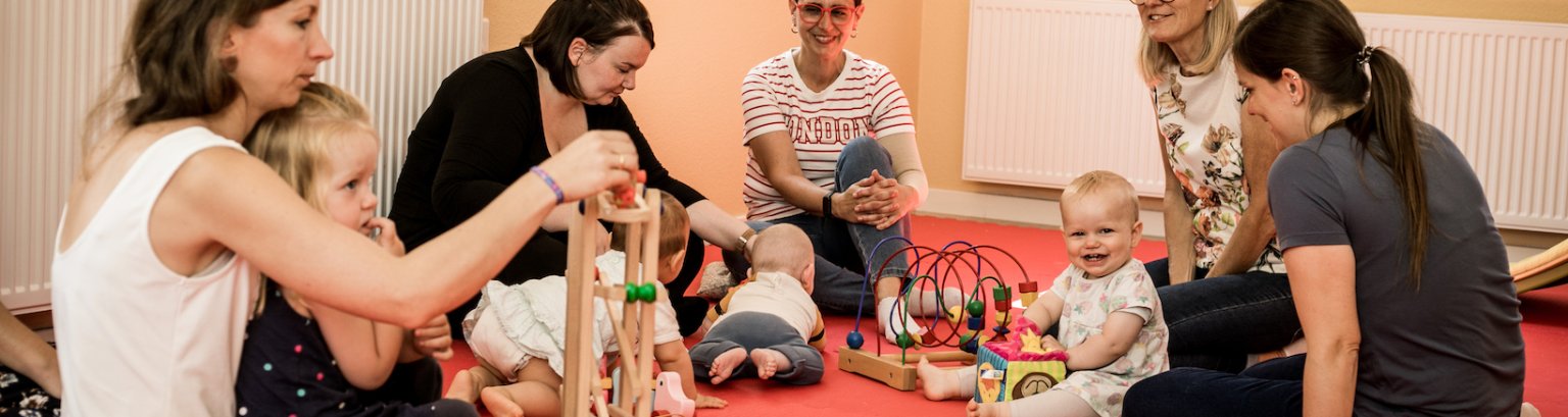 Bildausschnitt: spielende Kleinkinder auf einer Matte, die Mütter sitzen um sie herum, ihre Köpfe sind nicht zu sehen.
