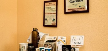 Ein Regal mit Tassen und anderen Gegenständen vor einer Gelben Wand. Dazu ein Bild mit der Aufschrift Tauschregal.