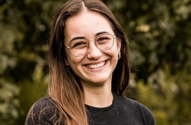 Eine junge Frau mit dunklen langen Haaren und Brille lächelt